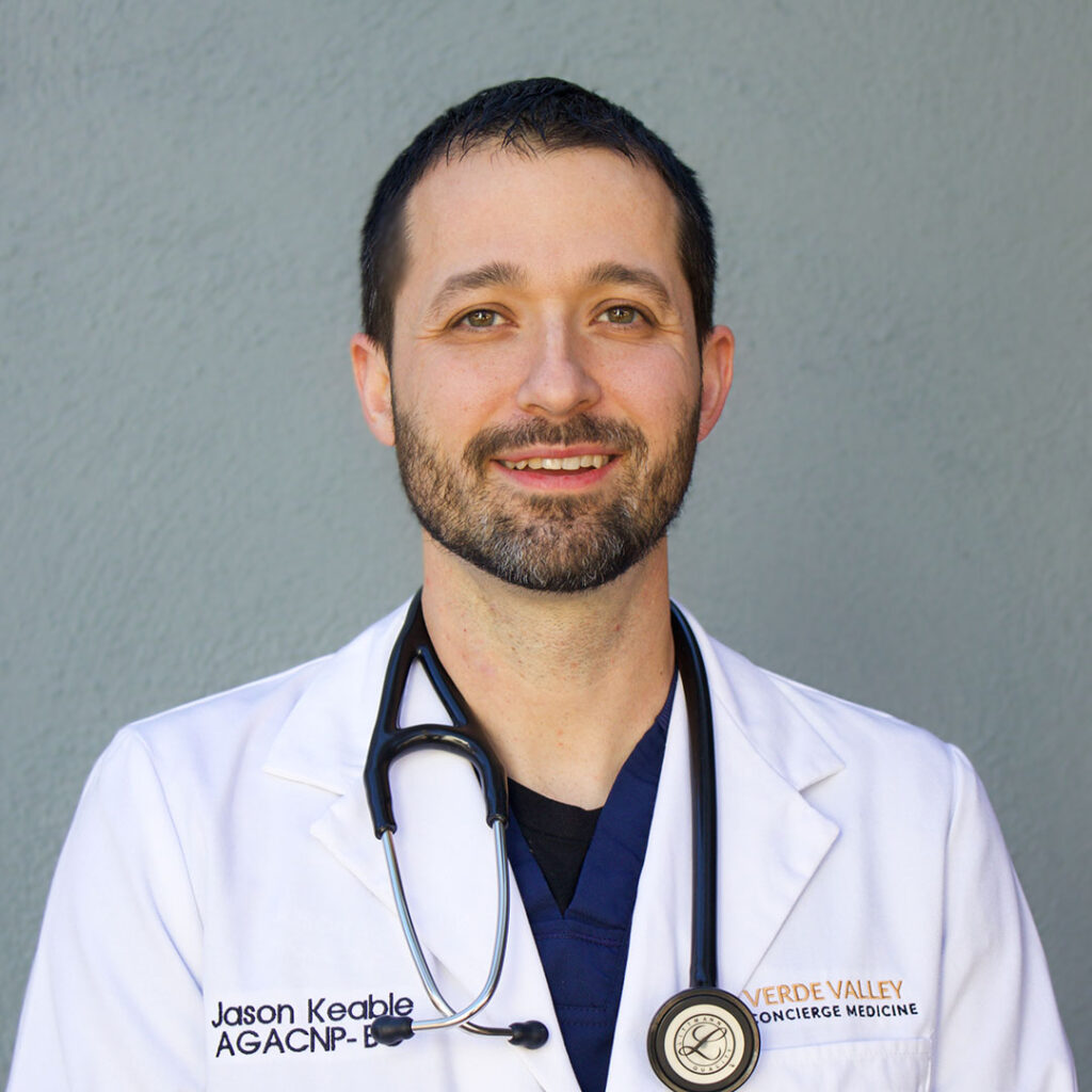 A man wearing a white coat with a stethoscope around his neck, posing against a gray background. The name "Jason Keable AGACNP-BC" is embroidered on his coat.