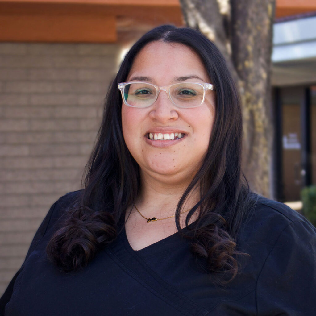 A woman with long dark hair, wearing glasses and a black shirt, stands outdoors smiling. A tree and building are in the background.