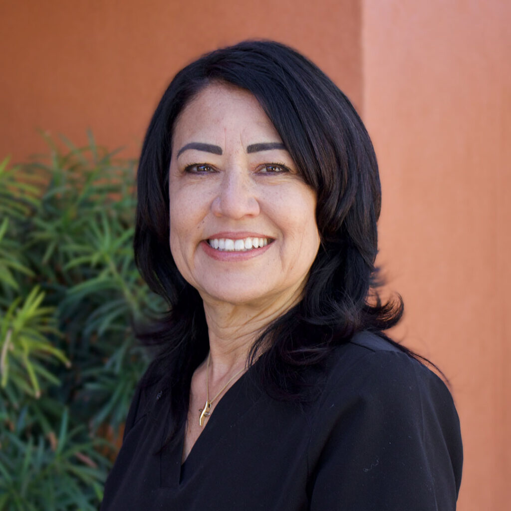 A woman with shoulder-length black hair and dark clothing smiles for a photo with green foliage and an orange wall in the background.