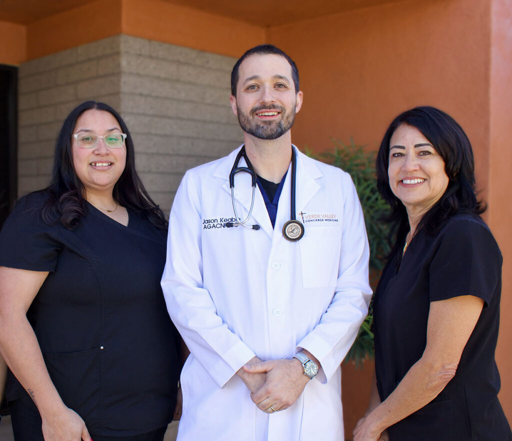 Three healthcare professionals, one man in a white coat with a stethoscope and two women in black scrubs, standing outside a building.